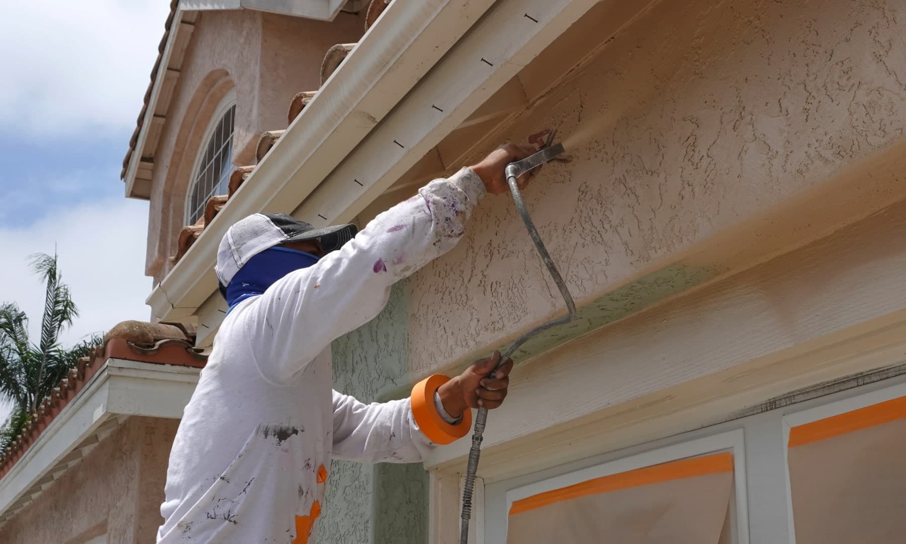 Painter spraying finish color along the fascia of a textured stucco exterior